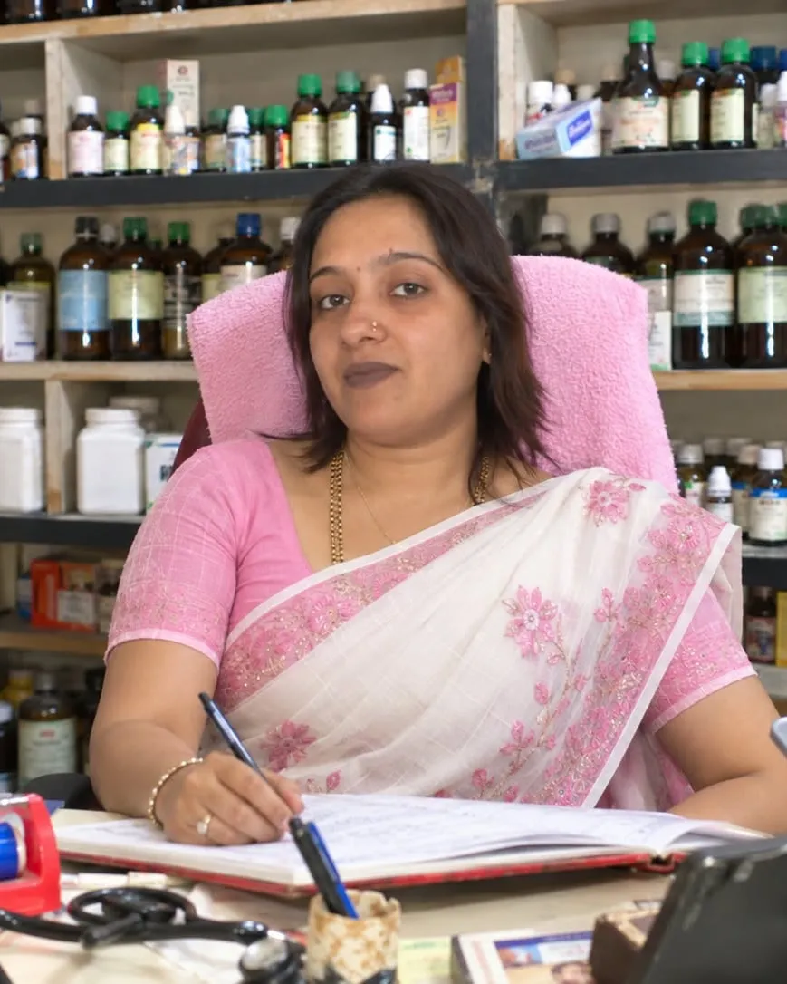 Dr. Sonal Jain at her desk in the clinic dispensary, writing in a register, with shelves of homoeopathic remedies behind her.